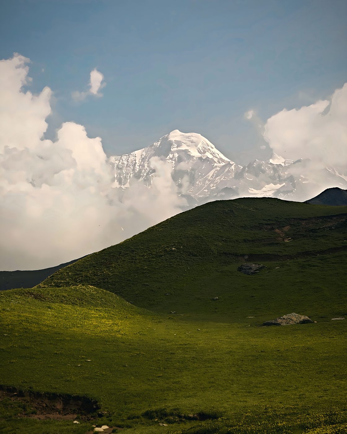A snow-capped peak rising out of clouds behind green rolling meadows