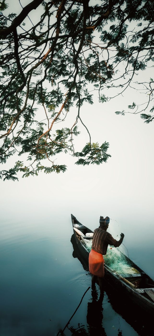 A fisherman in an orange mundu sorting nets beside his canoe in misty still water, under overhanging branches